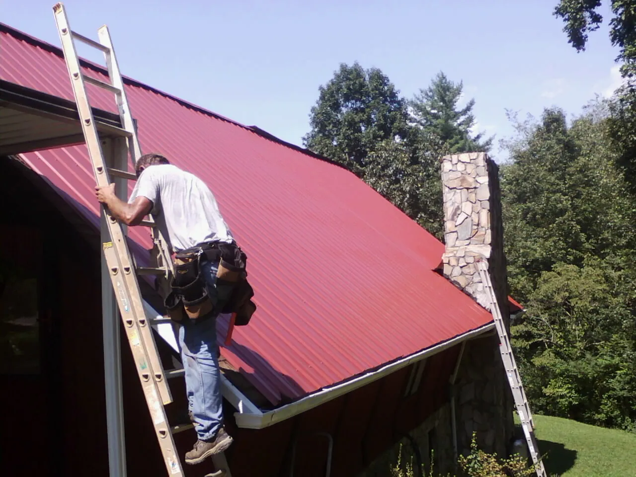 Roofer on a ladder beside a red metal roof and stone chimney — Soffit & Fascia Repair by Roofing Brothers in Corrales
